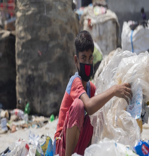 The image depicts a young boy wearing a red and black face mask, crouched down amidst a pile of plastic waste, collecting recyclable materials. Large sacks or bags of waste are visible in the background, suggesting a setting like a landfill, recycling area, or garbage dump. The image conveys the harsh reality of child labor in waste collection