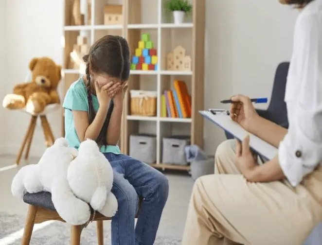 A sad girl sits holding a white teddy bear, her face in her hands. A counselor with a clipboard sits nearby. The brightly lit room has toys, books, and blocks, creating a child-friendly, comforting space.