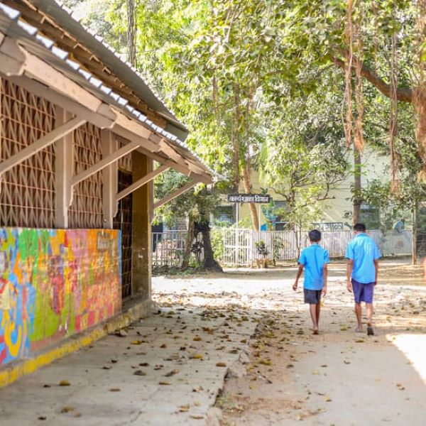 This image shows an outdoor institutional setting with a colorful building, open lattice walls, and a leafy pathway. Two children in blue uniforms walk toward a shaded area with trees and a distant fence.