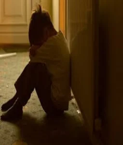 A child sitting on the floor, leaning against a wall, hugging their knees in a dimly lit room.