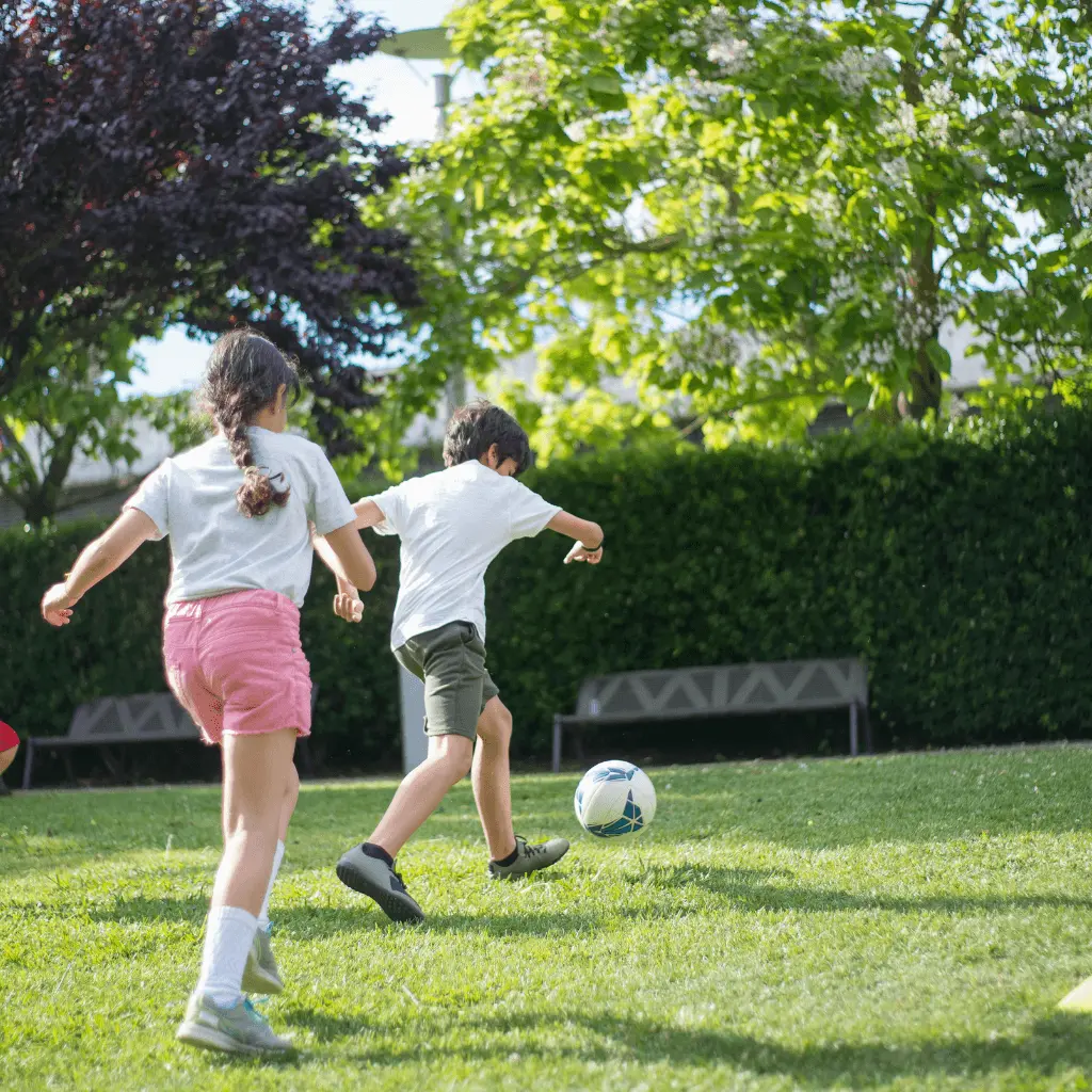 Four children are playing football outside on green grass, surrounded by trees. They look happy and active, enjoying the sunny day in an open park-like area.