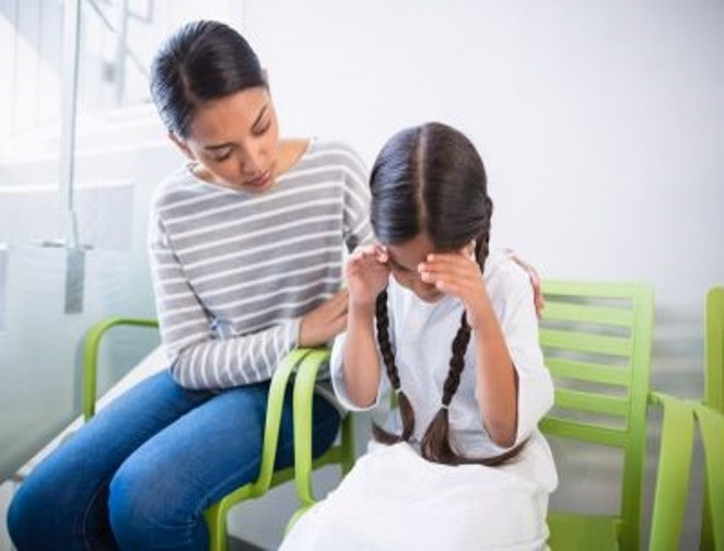 This image shows a young girl sitting on a green bench, looking upset with her hands on her face. Beside her, a woman in a striped shirt gently comforts her by touching her shoulder.