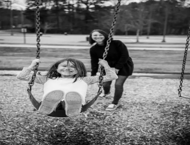 A child is swinging on a swing with a lady standing behind her who is pushing the swing in a playground