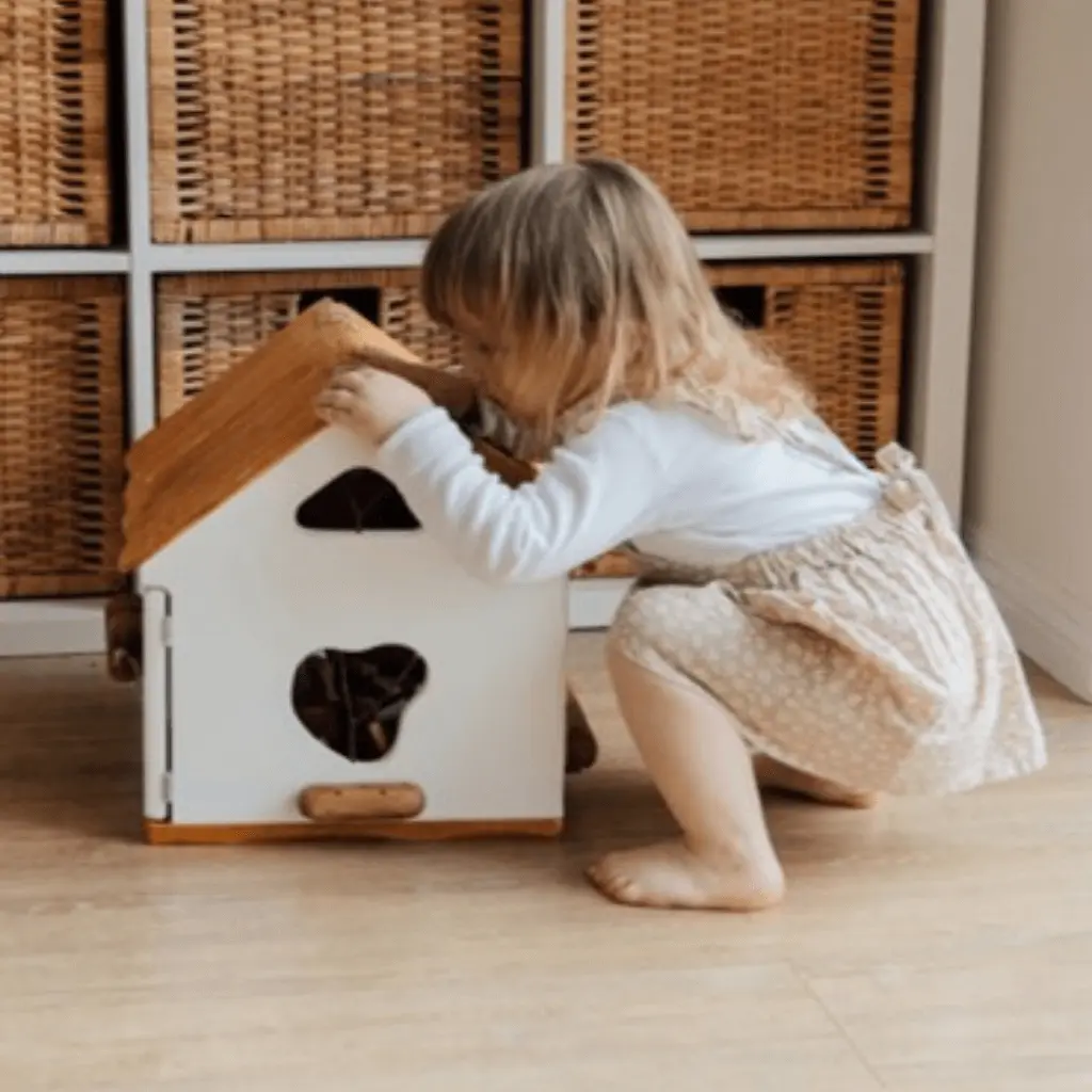 A toddler crouches on a wooden floor, curiously looking inside a small white dollhouse with a wooden roof, set in a cozy room with wicker baskets behind.