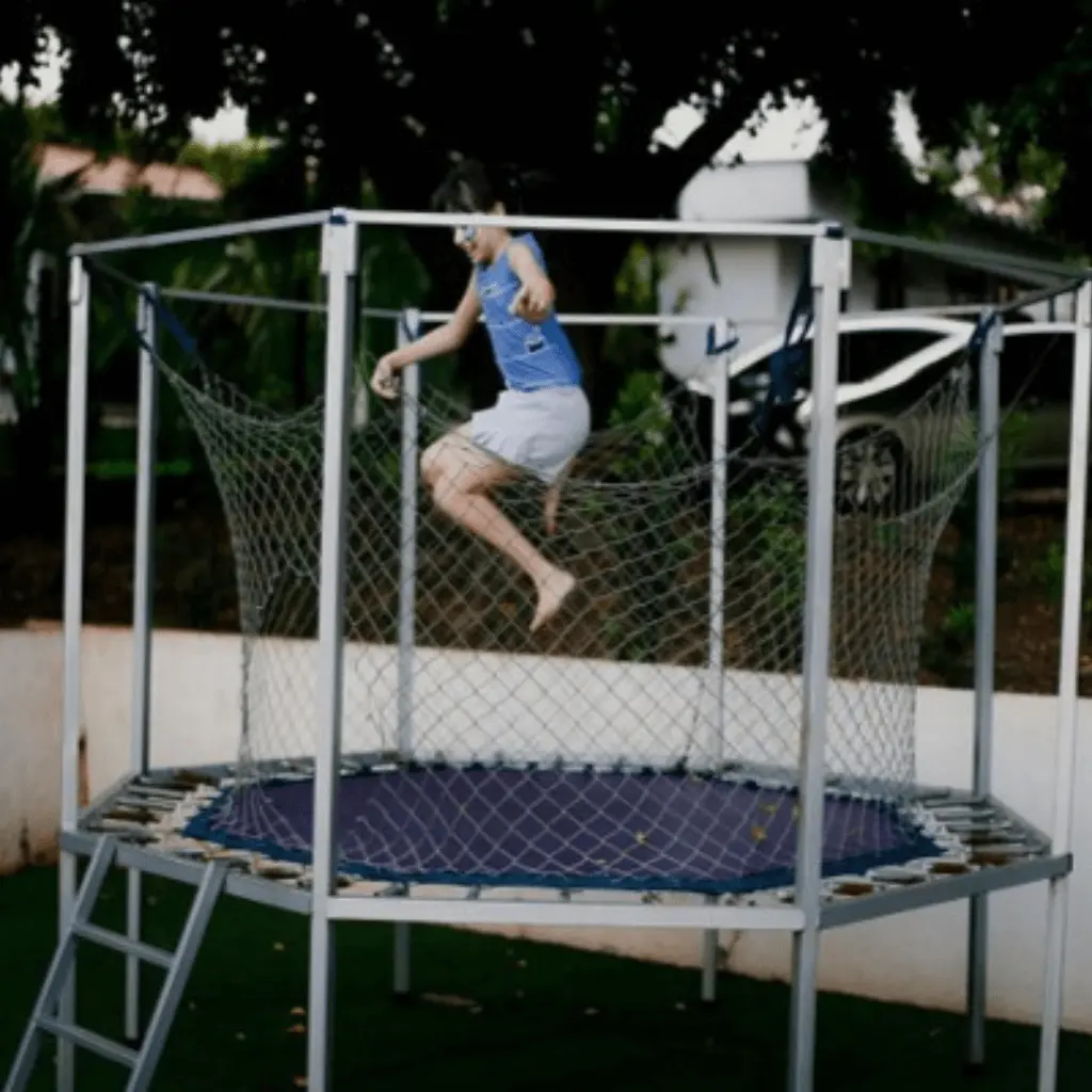 A child is jumping on a trampoline surrounded by a safety net. The trampoline has a metal frame and a ladder, with trees, a parked car, and a white wall in the background.